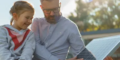 Un adult și o tânără fată inspectează un panou solar, zâmbind, în aer liber.