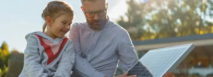 Un adult și o tânără fată inspectează un panou solar, zâmbind, în aer liber.