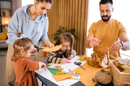 Egy nő levél alakú papírt vág, miközben két gyerek rajzol. Egy férfi néhány levelet tart a kezében.