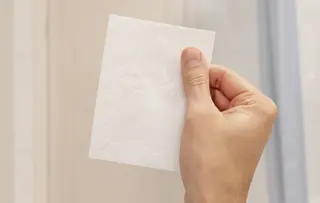 Eine Person hält ein Blatt Toilettenpapier vor sich, nur eine Hand ist sichtbar.